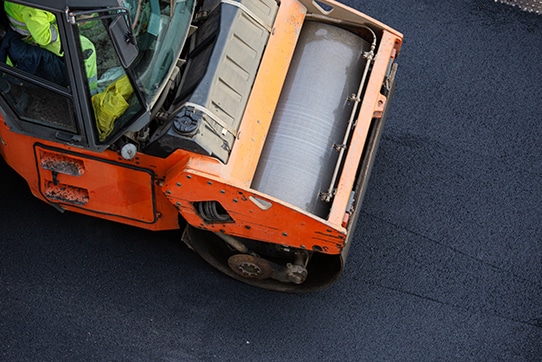 A construction vehicle with an orange steamroller compacts freshly laid asphalt on a road. View from above shows the steamrollers large cylindrical drum and part of the machinery.