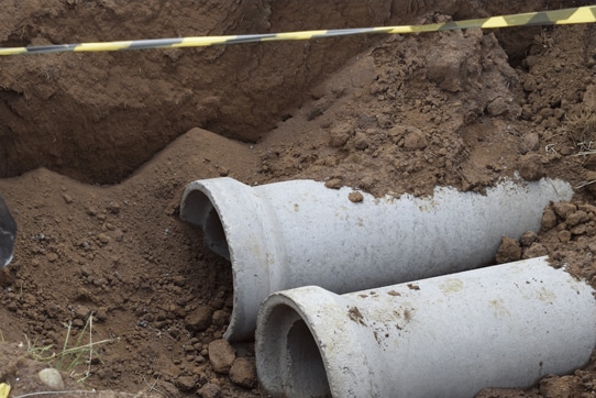 A partially dug trench with two large concrete pipes partially buried in soil. There is yellow and black caution tape above the trench. Dirt and earth are piled around the area.