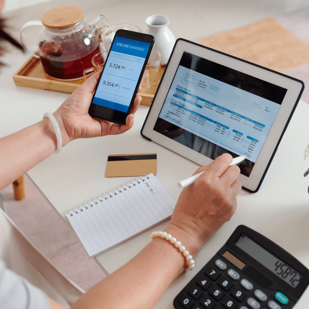 A person manages finances at a table, using a smartphone for online banking and a tablet displaying a financial report. A notepad, pen, calculator, credit card, and teapot with cups are also on the table.