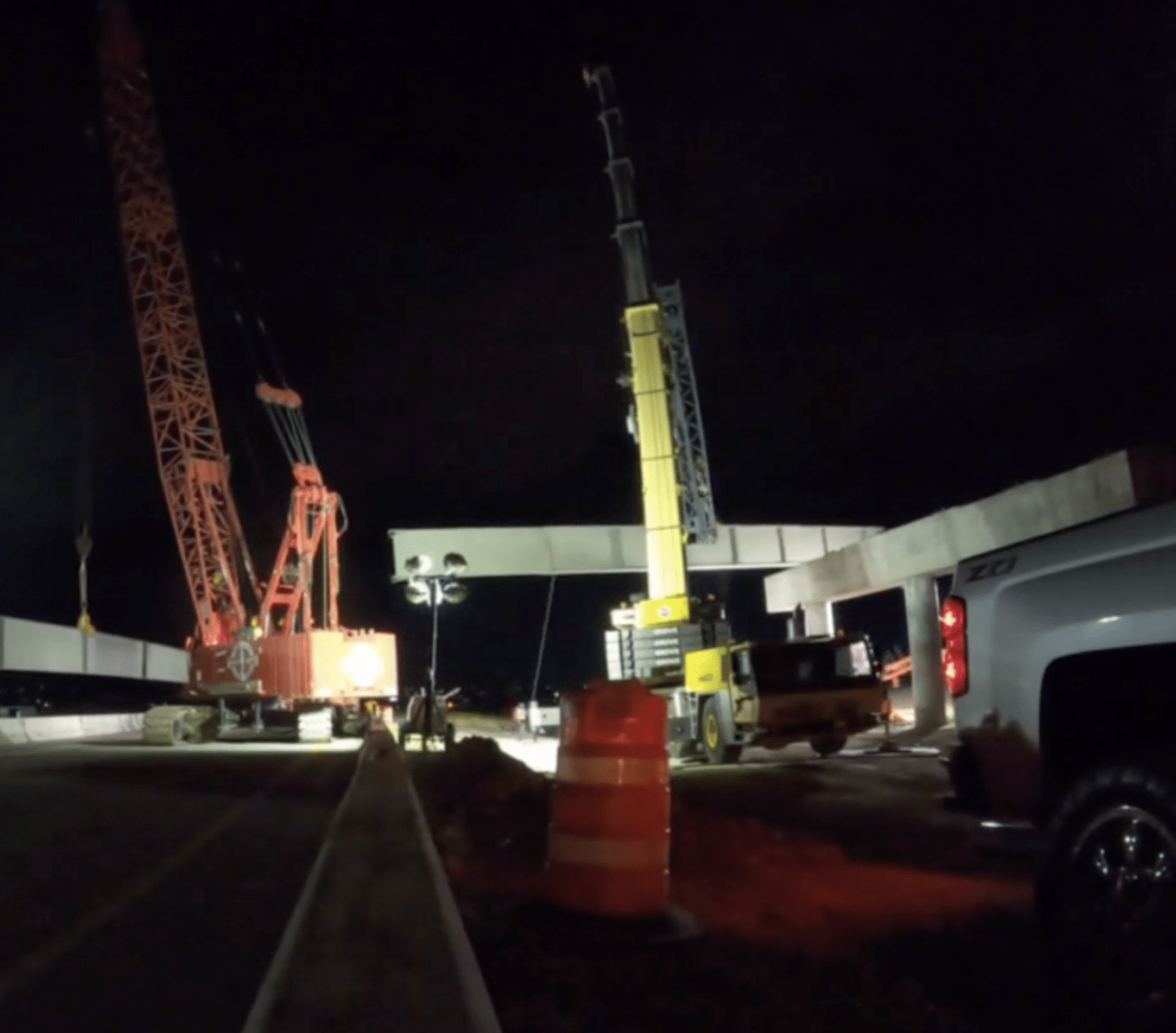 Night construction site with two large cranes lifting concrete beams. A white truck is parked nearby, and an orange traffic barrel is visible in the foreground. The scene is illuminated by portable lights.