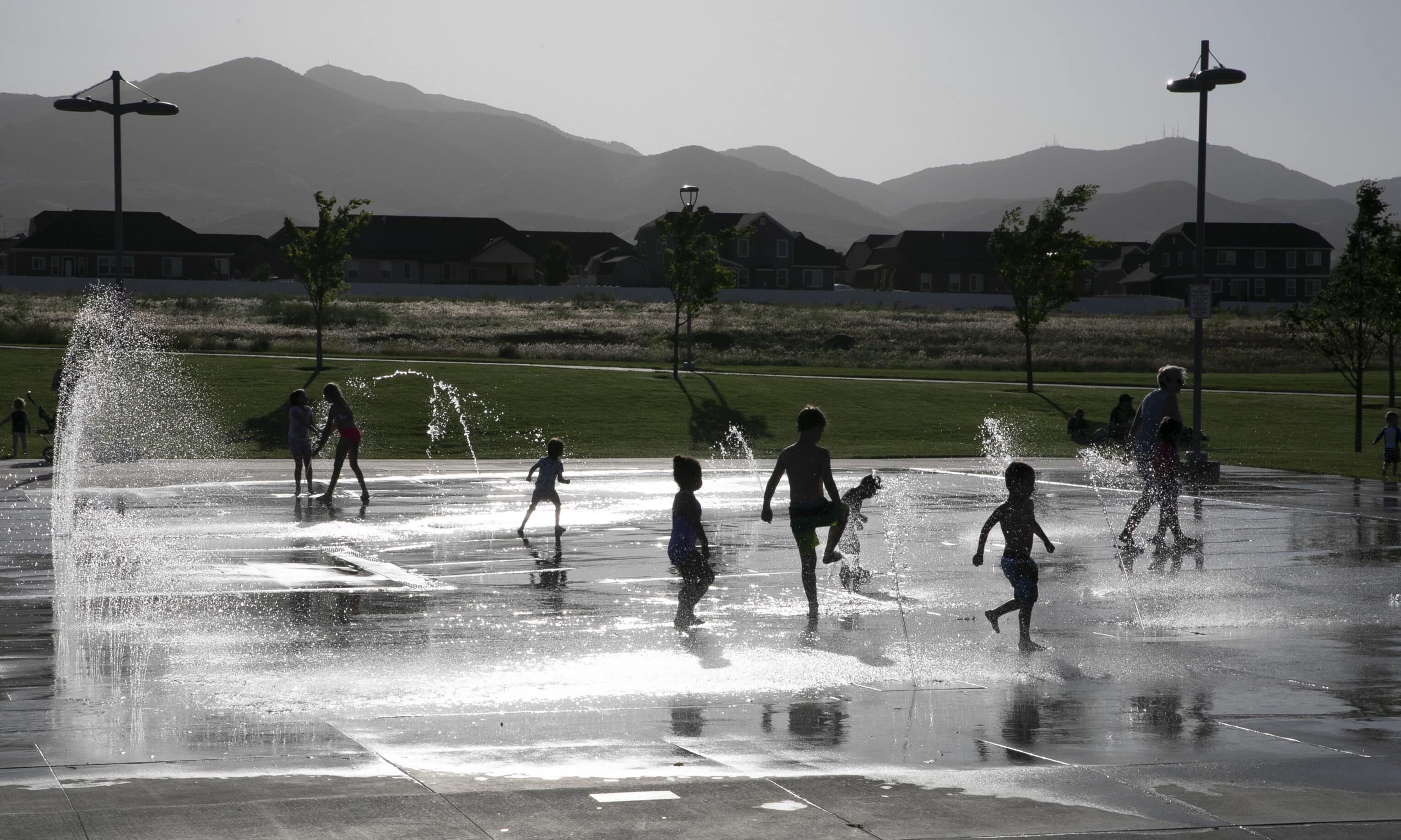 Children play and run through water fountains on a concrete plaza in a park at dusk. Shadows are cast as the sun sets behind distant mountains, with trees and buildings visible in the background.