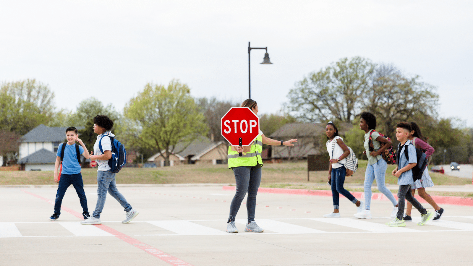 A crossing guard with a neon vest holds a stop sign, helping a group of children with backpacks cross the street at a crosswalk. The background shows houses and trees under a cloudy sky.