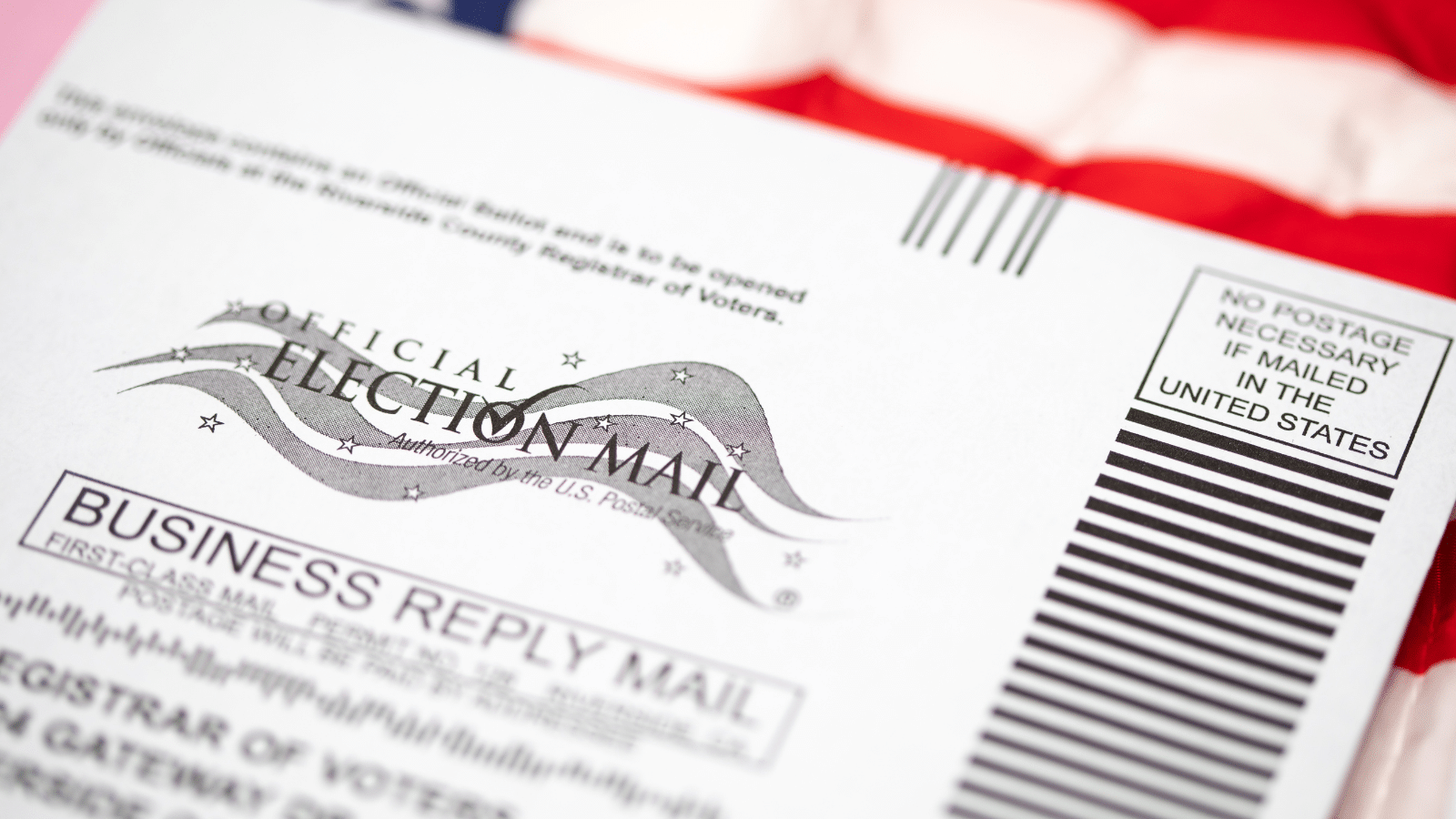 Close-up of an official election mail envelope with a business reply mail section. An American flag is partially visible in the background. The envelope states No postage necessary if mailed in the United States.