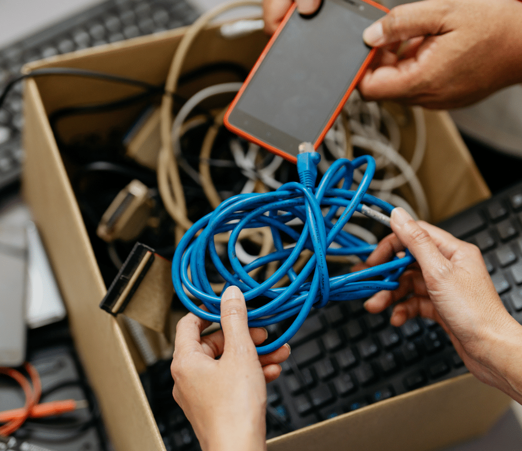 Two people sorting through a cardboard box filled with tangled electronic cables, a smartphone with a red case, and a keyboard. One person holds a bundle of blue cables, while the other holds the smartphone.