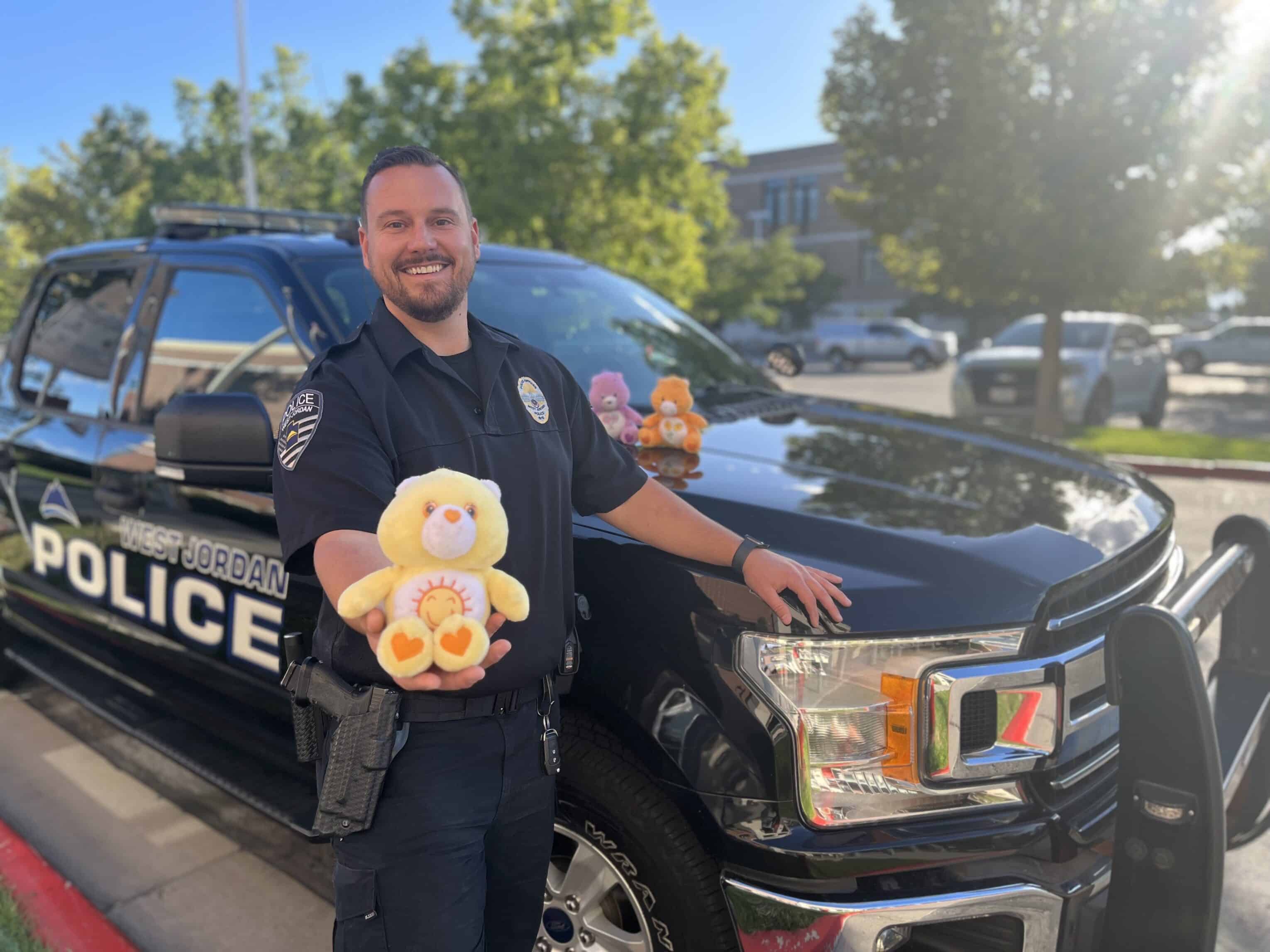 A police officer stands in front of a West Jordan Police vehicle, smiling and holding a yellow teddy bear. Three more teddy bears sit on the vehicles hood. Trees and a building are visible in the background.