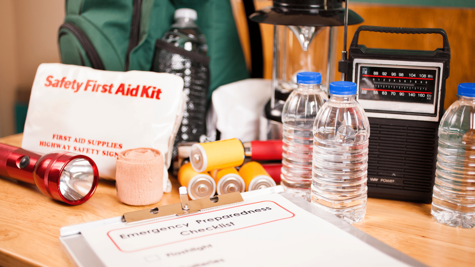 A collection of emergency preparedness supplies on a table, including a first aid kit, flashlight, water bottles, batteries, a hand-crank radio, and a lantern. A clipboard with an emergency preparedness checklist is also visible.