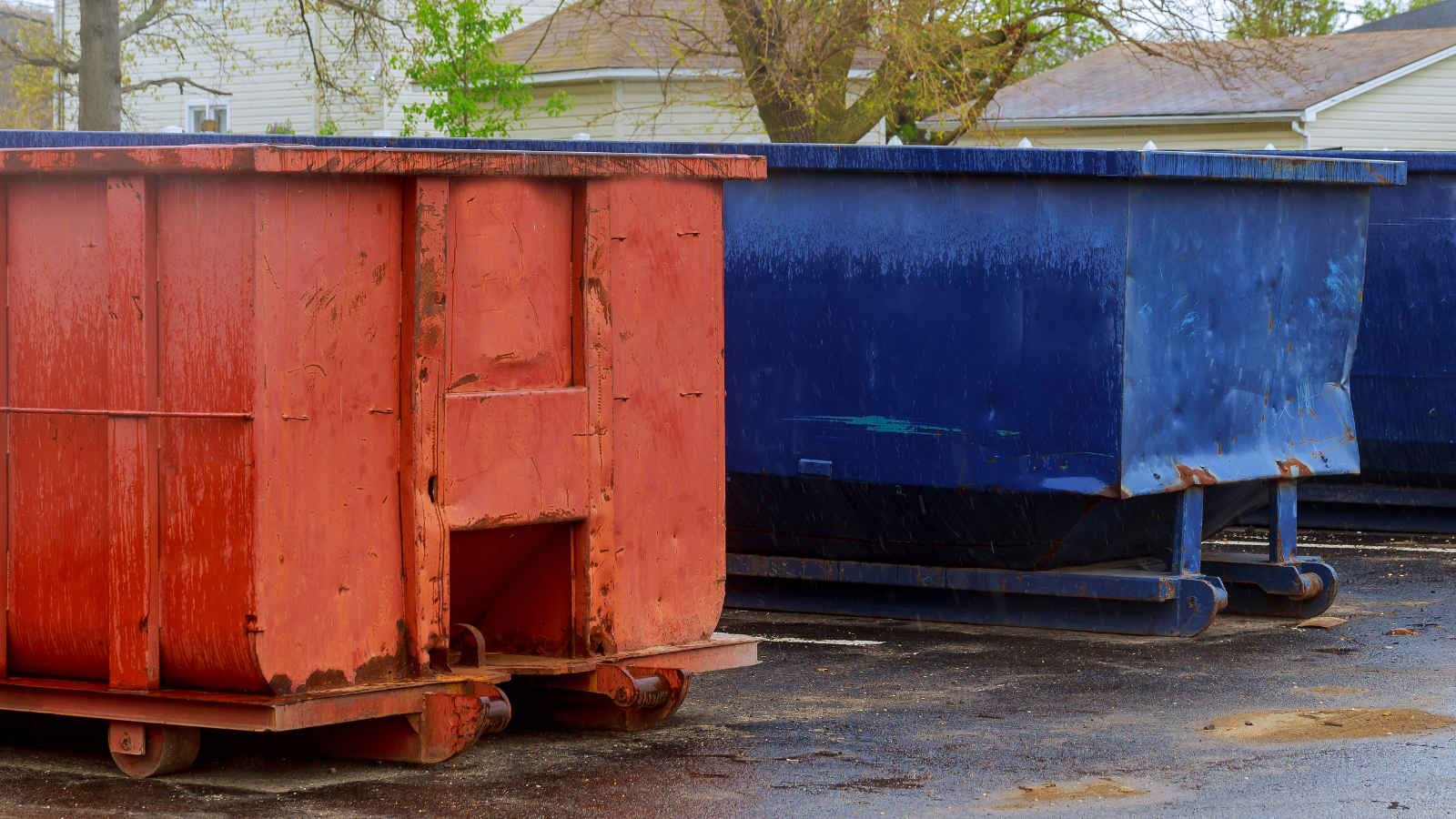 A red dumpster stands on the left next to a blue dumpster on the right, positioned on a wet asphalt surface in an outdoor area with trees and buildings in the background.