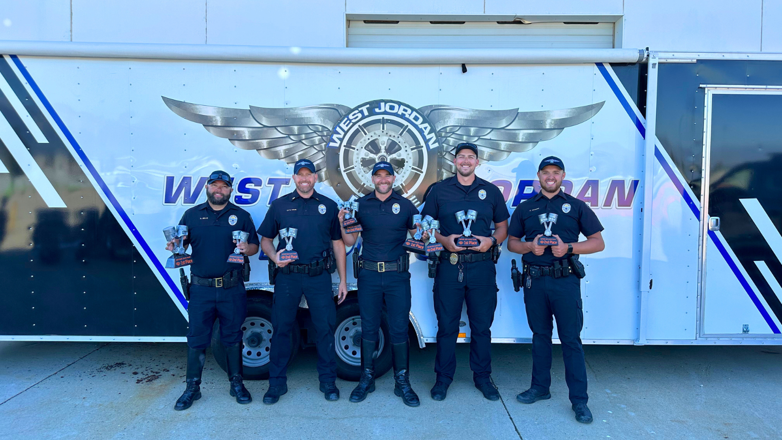 Five uniformed police officers stand smiling, each holding a trophy, in front of a trailer with the words West Jordan and a winged logo.