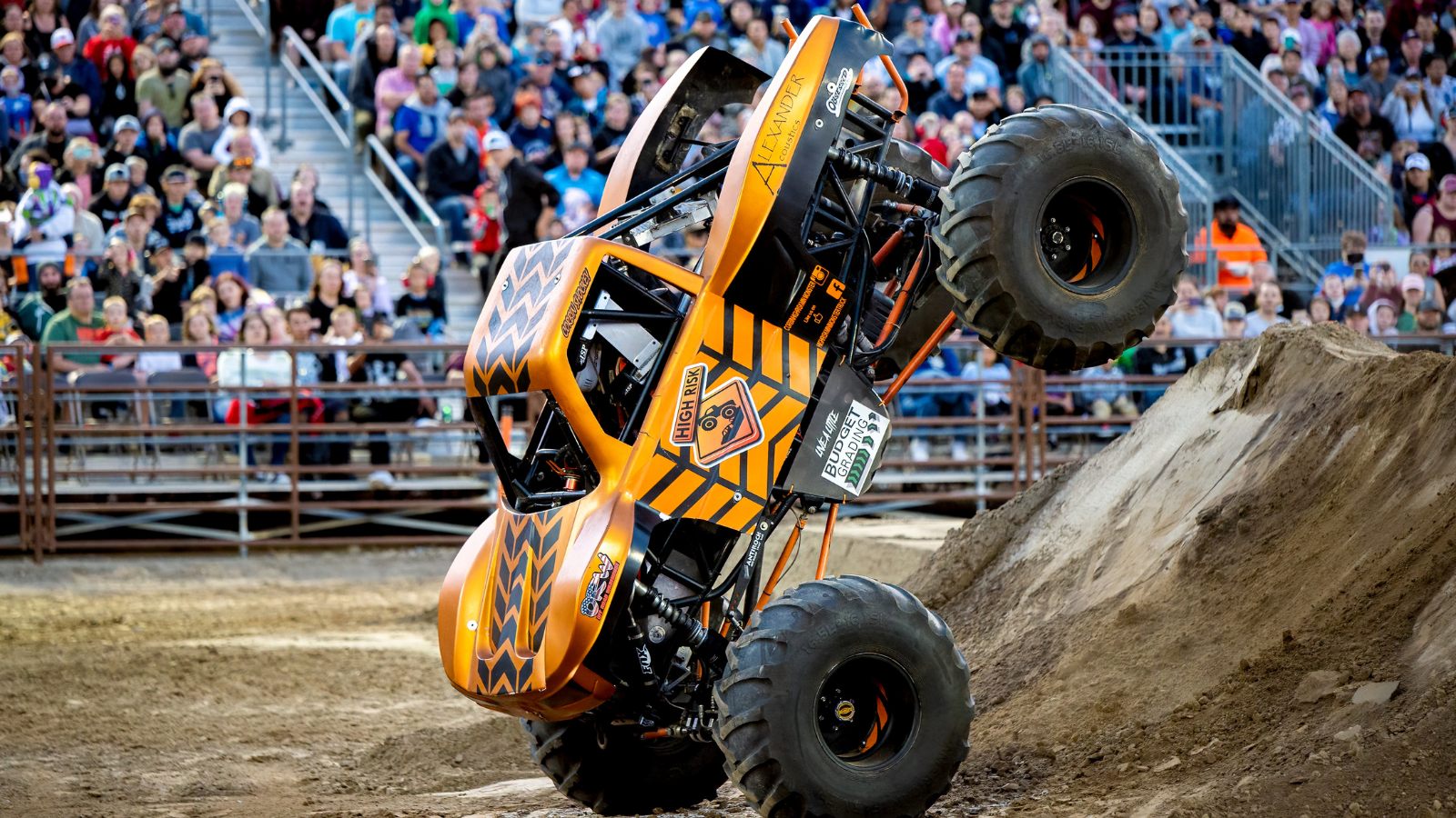 A bright orange monster truck with a zigzag pattern performs a wheelie on a dirt ramp in a stadium filled with spectators. The trucks large tires and elevated position emphasize its power and agility.