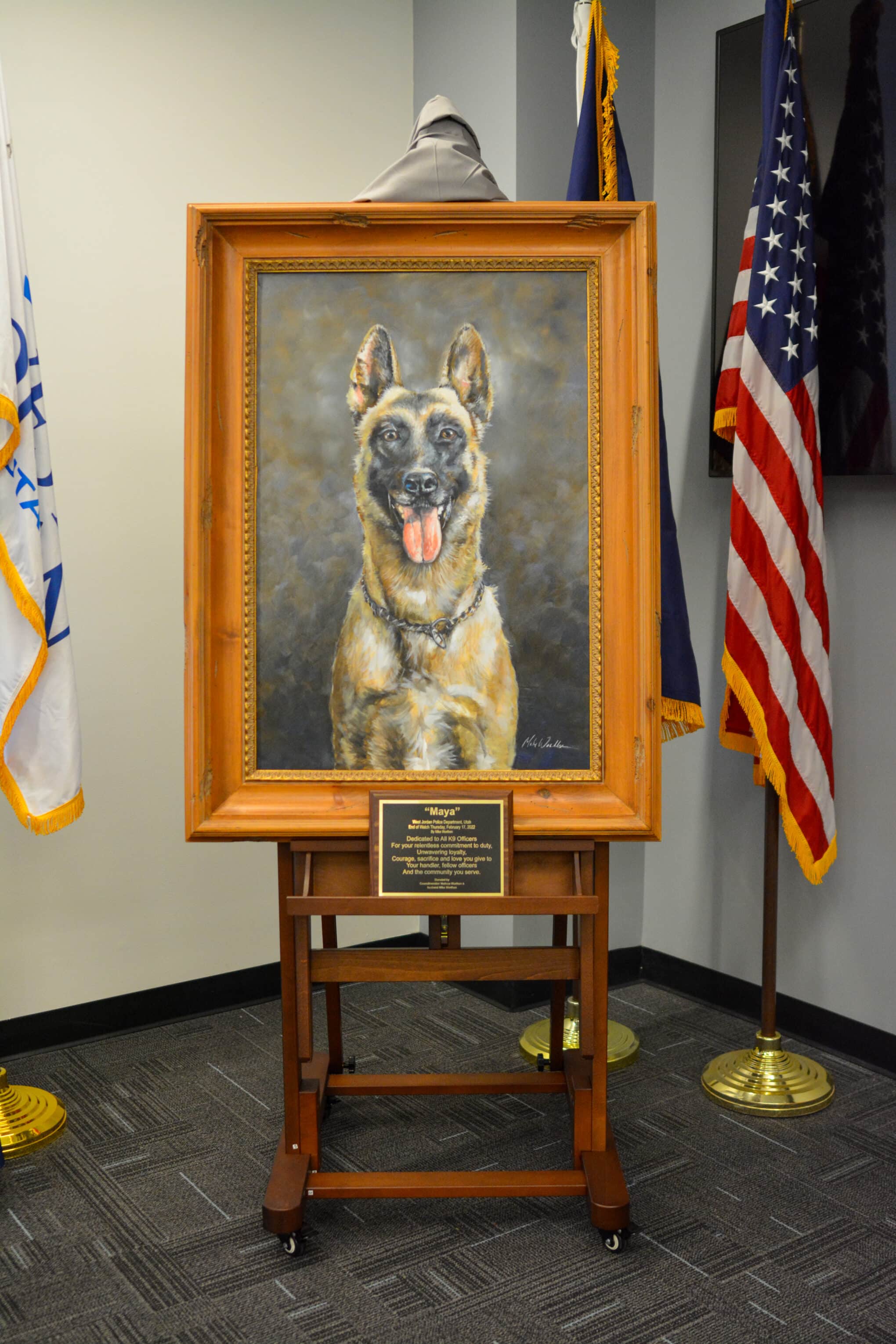 A framed portrait of a German Shepherd with its mouth open is displayed on an easel. The plaque below the painting reads Above and Beyond. American flags are visible on the right side.