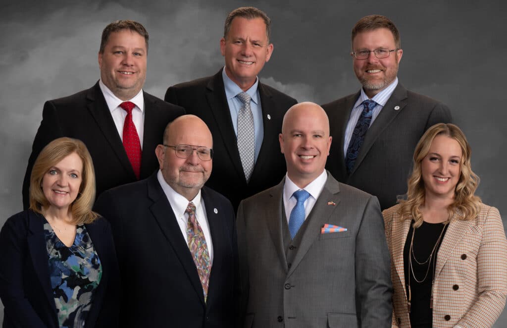 A group of seven people, consisting of five men and two women, dressed in formal business attire, posing together against a gray background. Some are wearing suits and ties, while others have blazers. They all smile towards the camera.