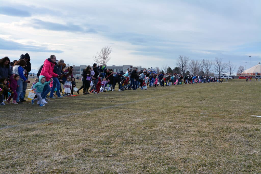 A large group of people, including children holding baskets, stand in a line on a grassy field. They appear to be preparing for an event, possibly an egg hunt. The sky is overcast, and some bare trees and distant buildings are visible.