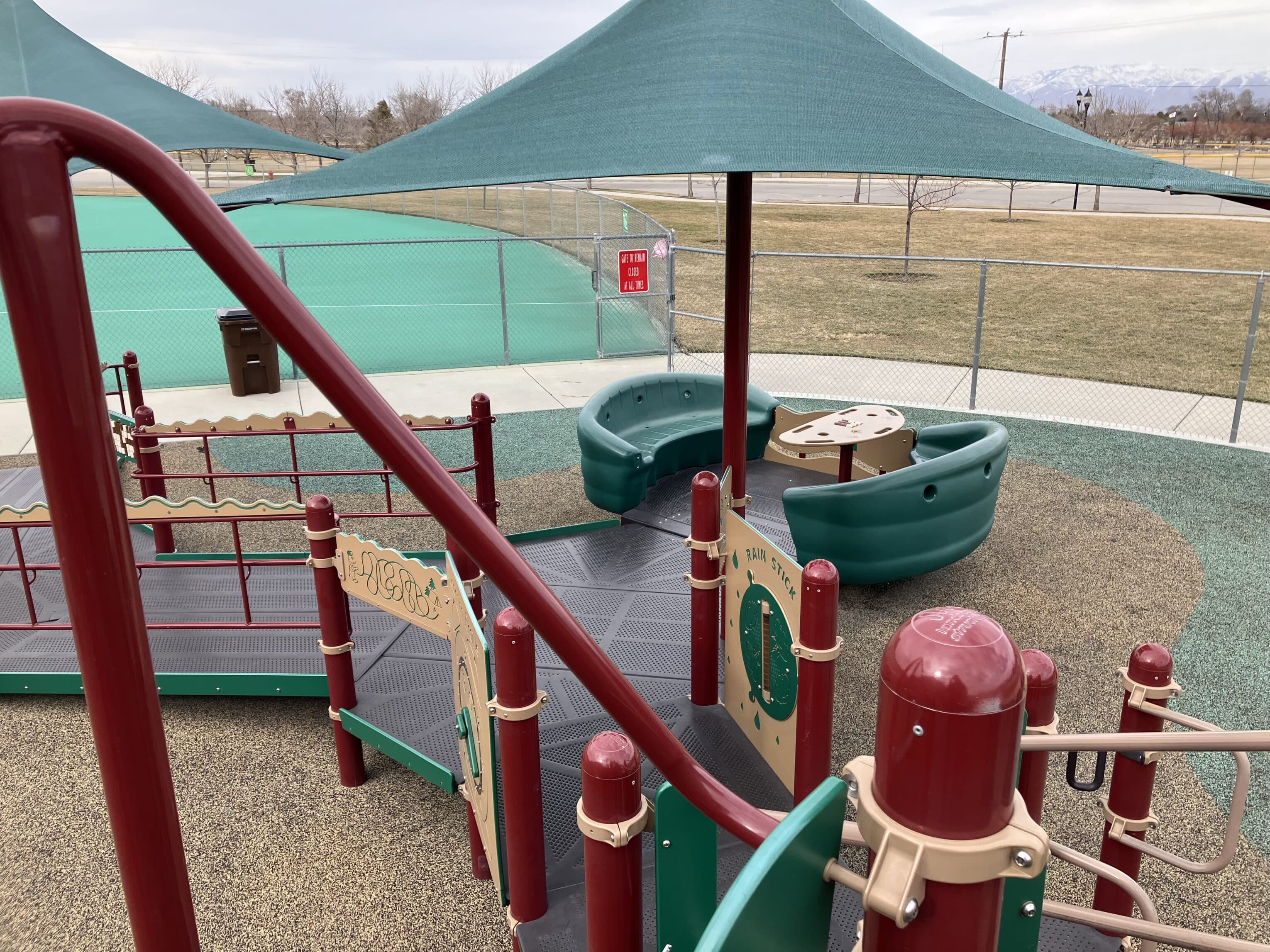 Playground area with accessible equipment, including a ramp, sensory panels, and green shade structures. The ground is covered with rubber safety surfacing. Theres a fence surrounding the playground and a grassy field in the background.