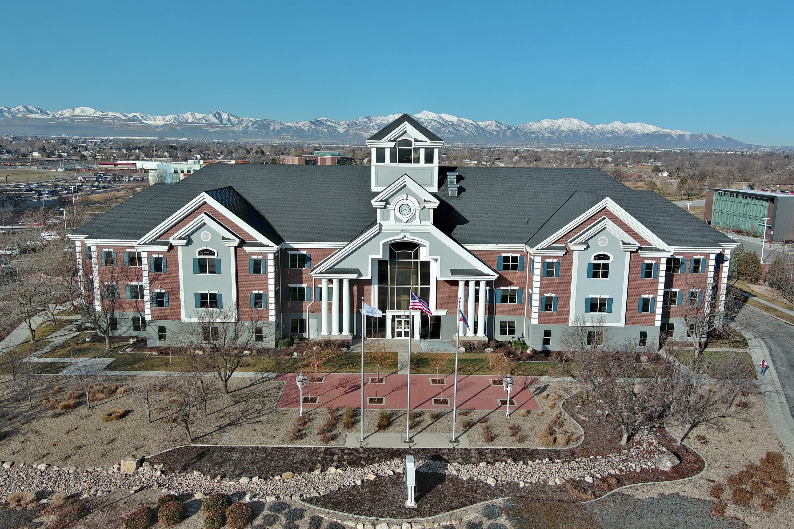 Aerial view of a large, red-brick building with white accents and a central clock tower. American flag flying in front. Snow-capped mountains in the background under a clear blue sky. Surrounding landscaped grounds with trees and pathways.