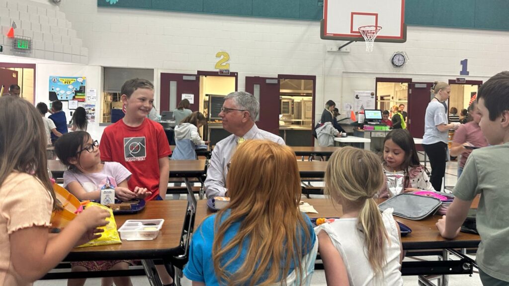 A group of children sits around a lunch table in a school cafeteria, chatting with an adult male. A basketball hoop is visible in the background, and cafeteria workers are seen behind a counter. The atmosphere is lively and friendly.