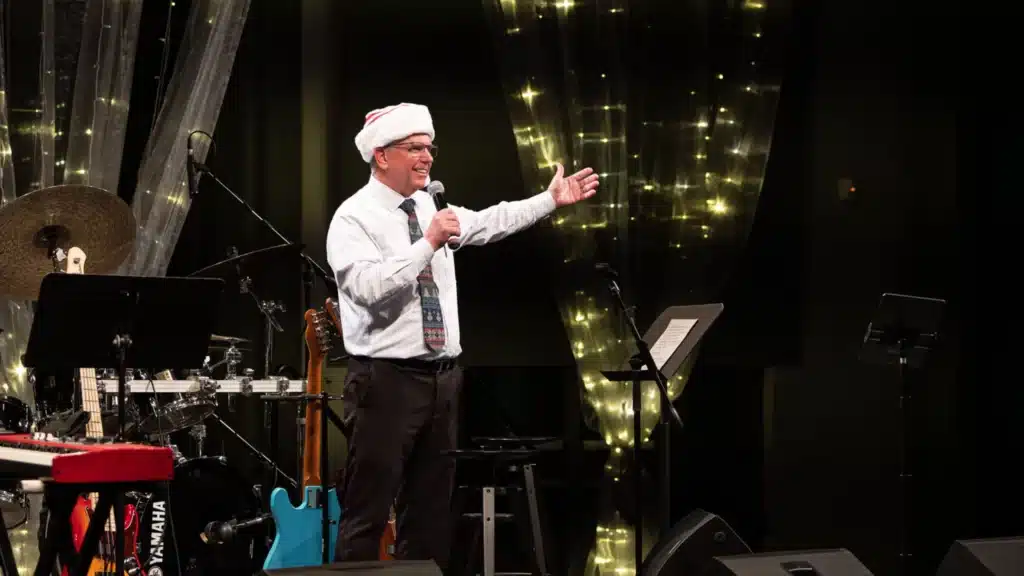 A man wearing a Santa hat and tie is speaking into a microphone on a stage decorated with string lights. Musical instruments, including a drum set and a blue electric guitar, are visible in the background. It appears to be a festive event.
