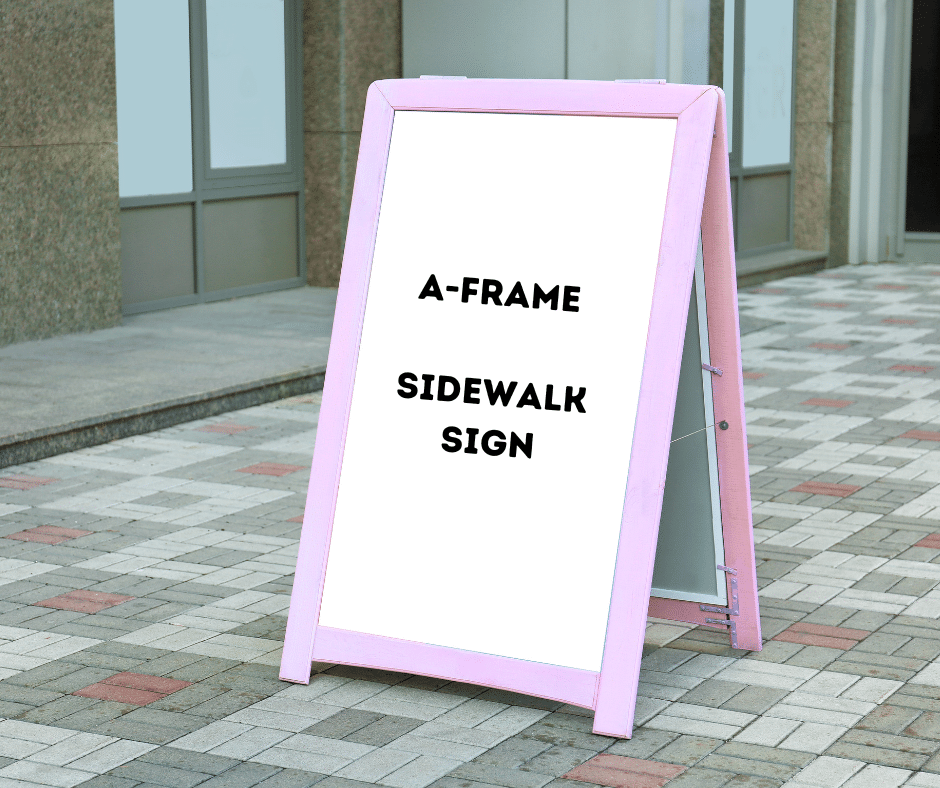 A pink A-frame sidewalk sign with the text A-FRAME SIDEWALK SIGN stands on a paved walkway in front of a building with large windows.