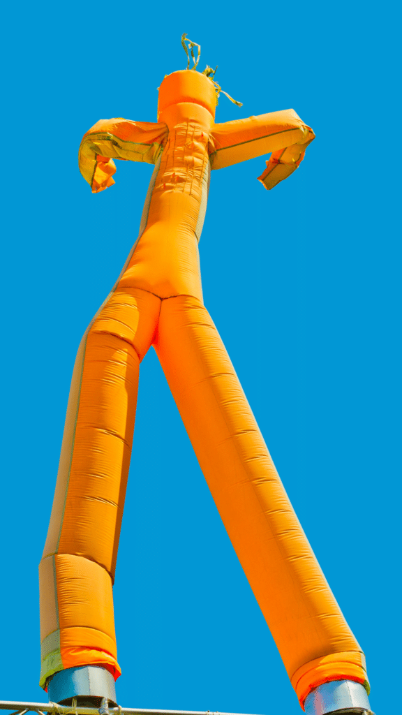 A tall, orange inflatable tube man with outstretched arms moves in the wind against a clear blue sky.