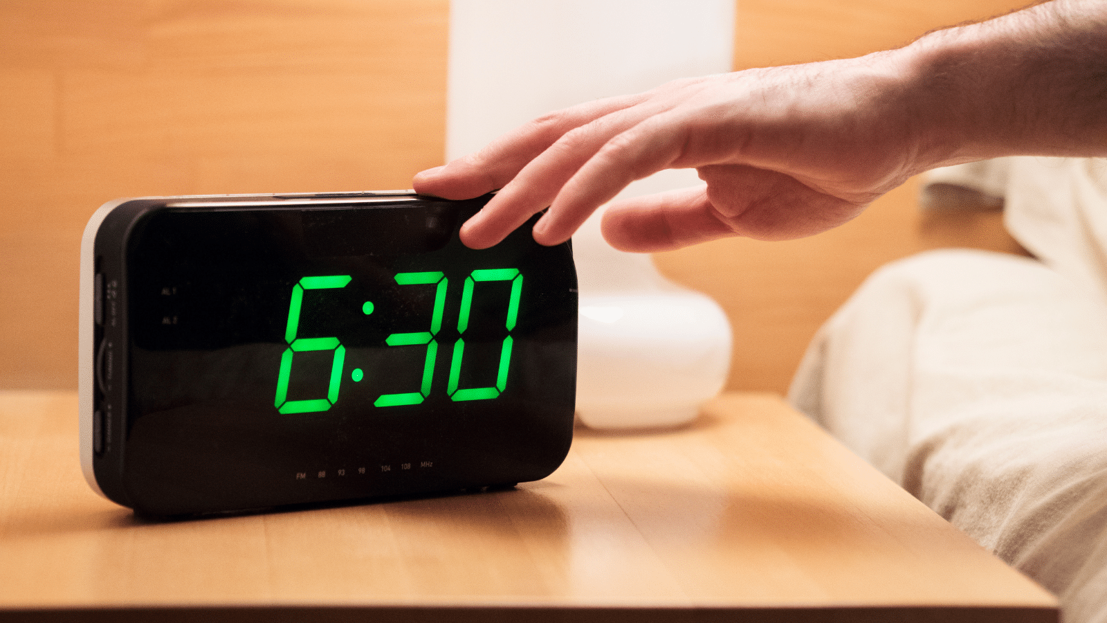 A hand reaches out to an electronic alarm clock displaying 6:30 in bright green numbers, placed on a wooden bedside table next to a white lamp.