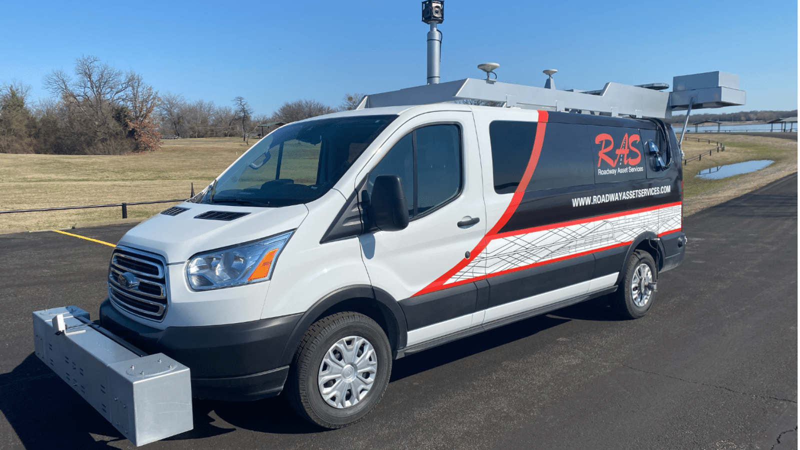 A white van with the logo R2S and website www.roadsafesystems.com on its side. The van has a large sensor and equipment mounted on top and a rectangular structure attached to the front. It is parked on an asphalt road near grassy fields.
