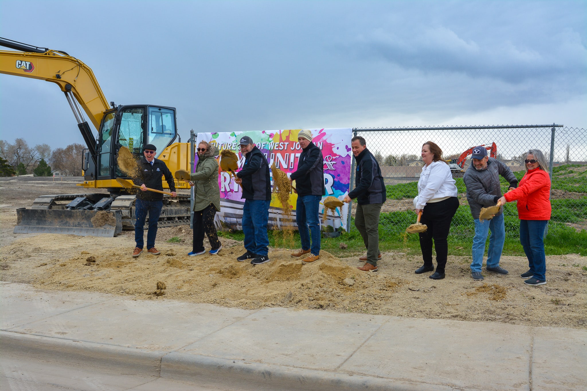 A group of eight people stands in a line, holding shovels and tossing dirt, participating in a groundbreaking ceremony. They are in front of a construction site with a banner in the background and a large excavator beside them.