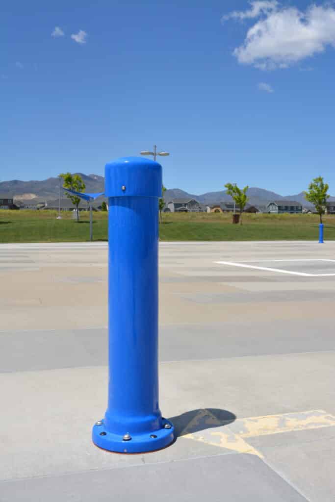 A tall, bright blue bollard stands on a large, empty concrete surface under a clear blue sky. In the background, there are distant houses, trees, and mountains.