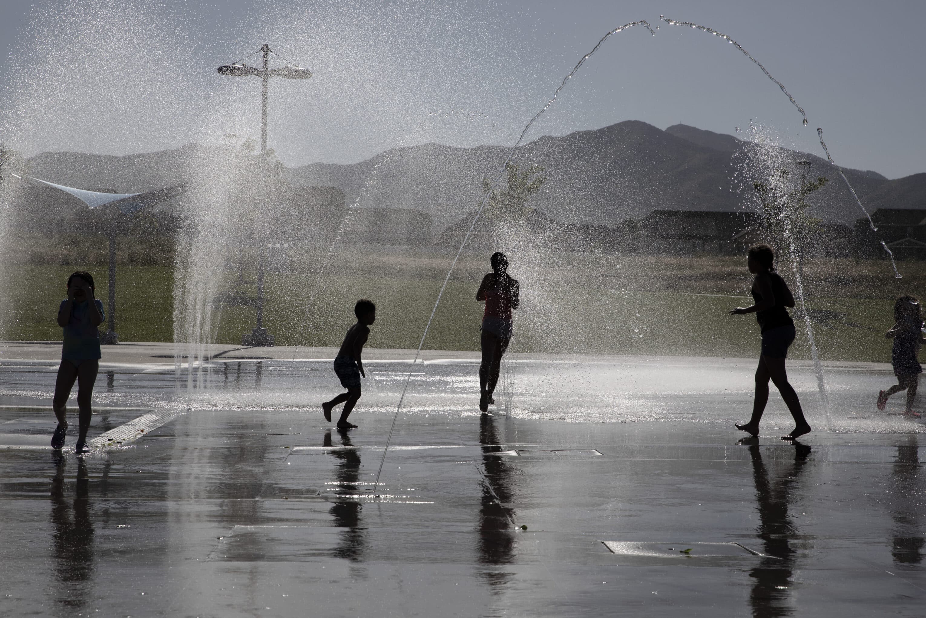 Children play in a splash pad, silhouetted against the sunlight. Water jets arch above them, creating a playful scene. Mountains and a grassy field are visible in the background under a clear sky.