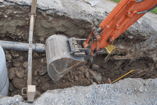 An excavator with an orange arm and a metal bucket is digging into the ground at a construction site. The trench is filled with rocks and dirt, and tubes run alongside it. A round concrete pipe is visible on the left side.