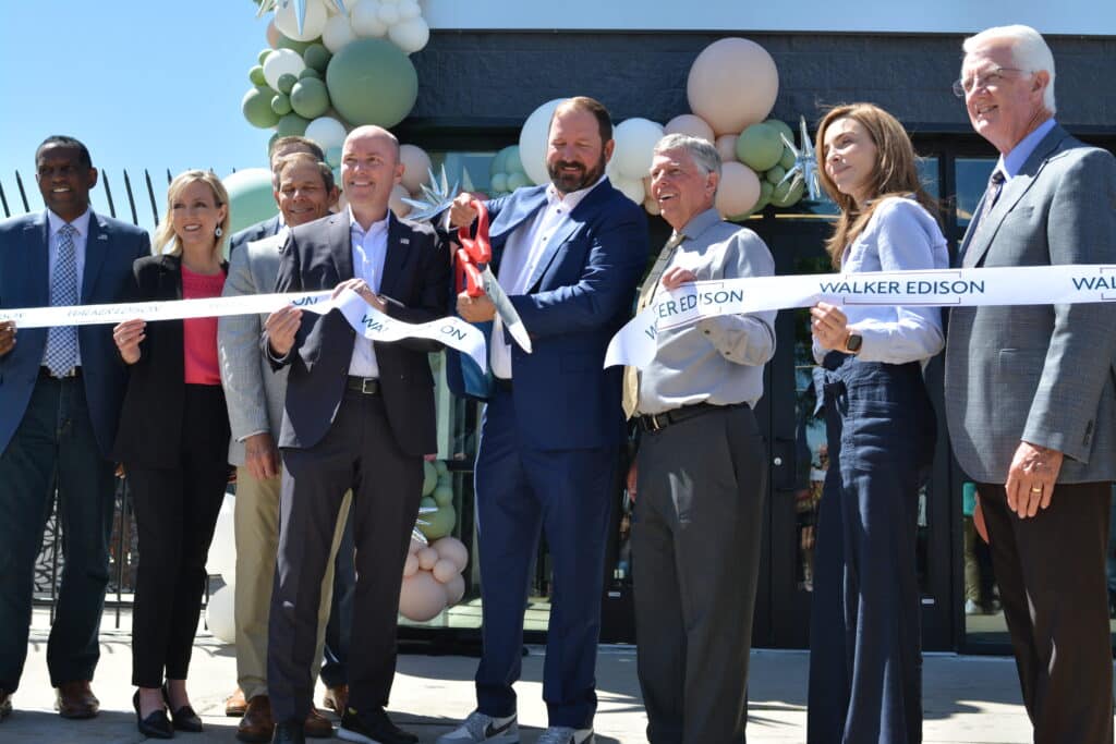 A group of people stand in front of a building adorned with balloons, smiling as a man cuts a ribbon with large scissors. The ribbon reads Walker Edison.