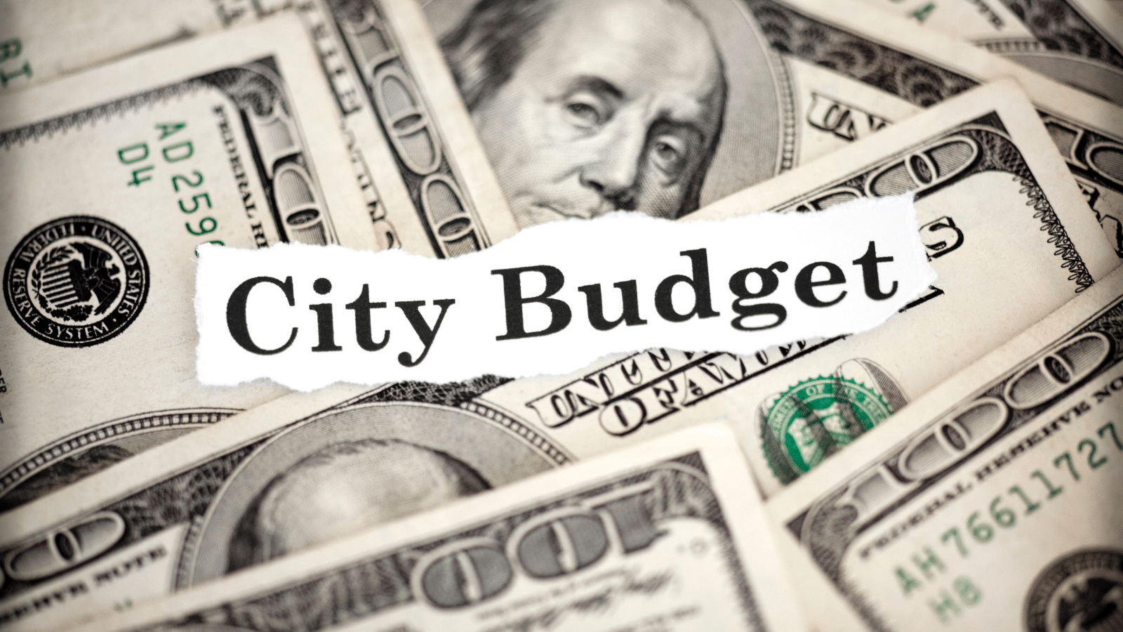 A close-up image of multiple U.S. one hundred dollar bills with a torn piece of paper on top. The paper has the words City Budget typed on it, suggesting a theme of financial planning or municipal budgeting.