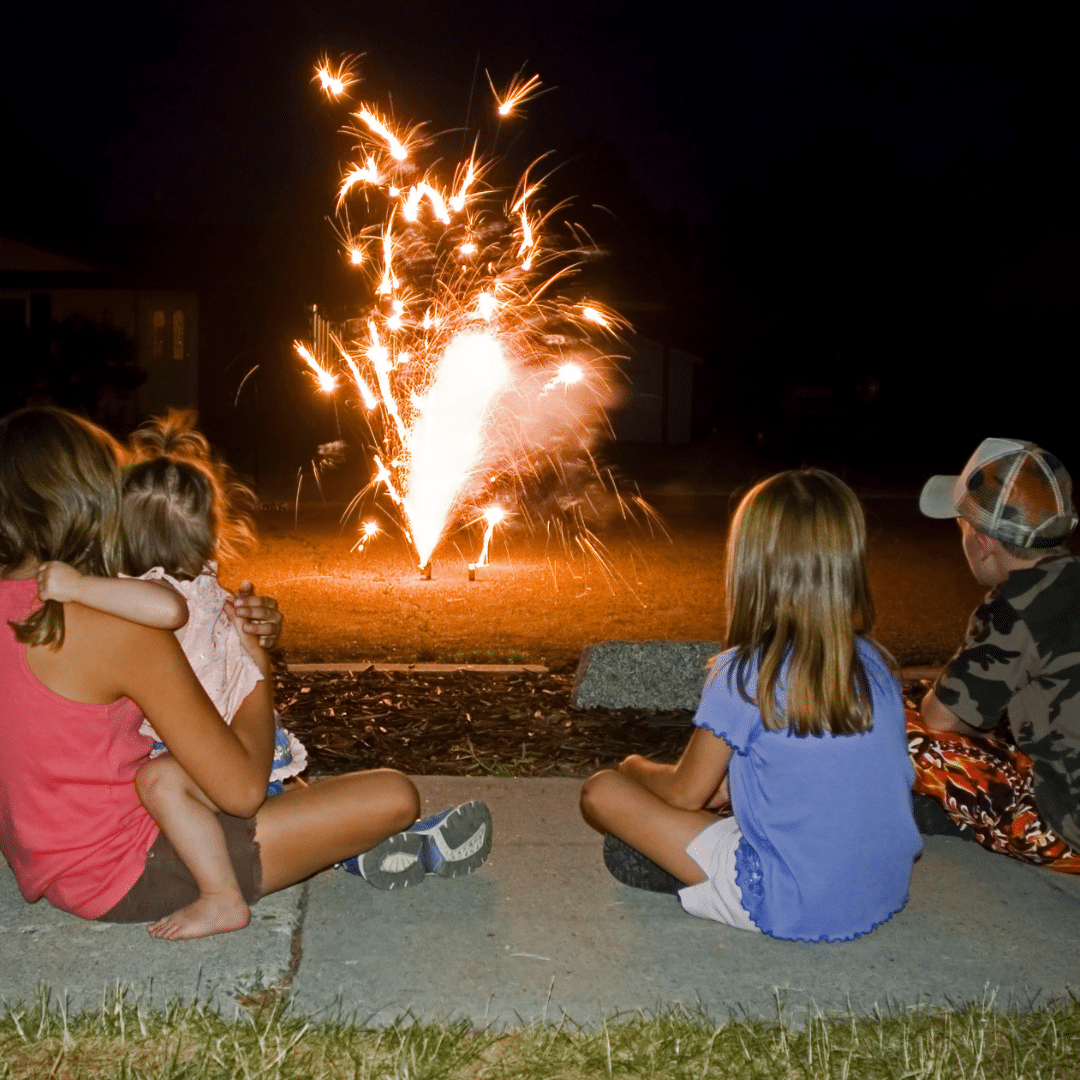 Four children sit on a sidewalk at night, watching fireworks in the distance. Two girls sit on the left, one holding a smaller child. A girl in a blue shirt and a boy in a cap sit on the right. Vibrant sparks illuminate the dark background.