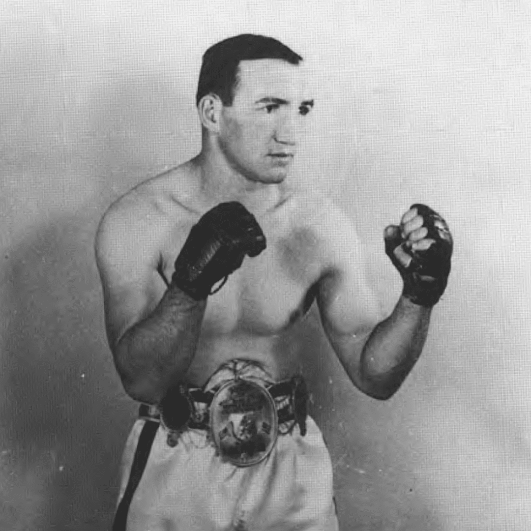 A vintage black-and-white photo of a boxer in a fighting stance, wearing boxing gloves and a championship belt. The boxer is shirtless and looks focused, with his fists raised and eyeing an opponent.
