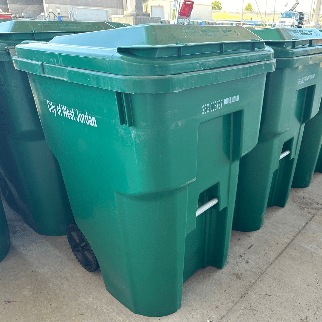 A row of green garbage bins with wheels, labeled City of West Jordan, is lined up on a concrete surface. The bins have white serial numbers and are partially overshadowed by a nearby building.