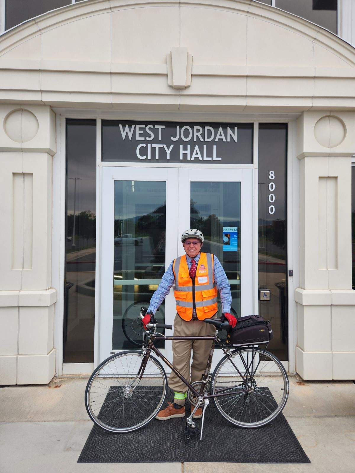 A person wearing a reflective orange vest and bike helmet stands with a bicycle in front of West Jordan City Hall. The building has glass doors, light-colored walls, and the address 8000 is visible.