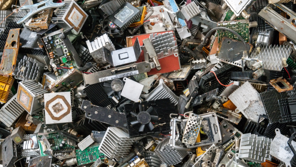 A pile of discarded electronic components, including circuit boards, fans, and various metal and plastic parts, mixed together in a heap. The components show signs of wear and usage.