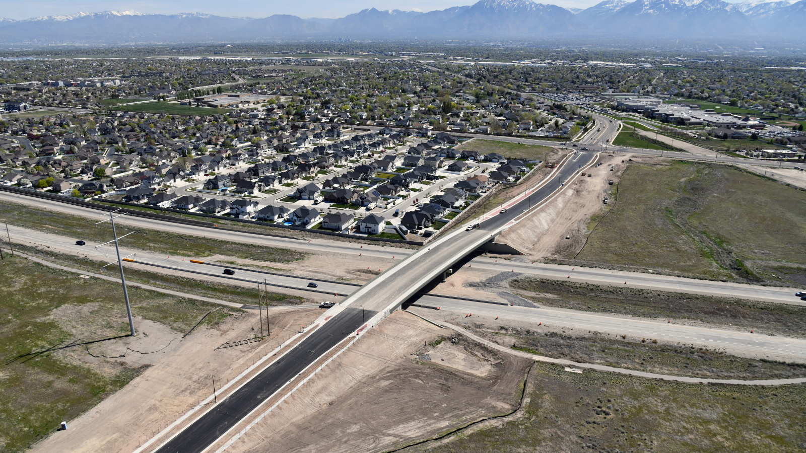 Aerial view of a newly constructed road and bridge intersecting a suburban neighborhood with houses, surrounded by open fields. Vehicles can be seen on the roads, with a backdrop of mountains under a clear sky.