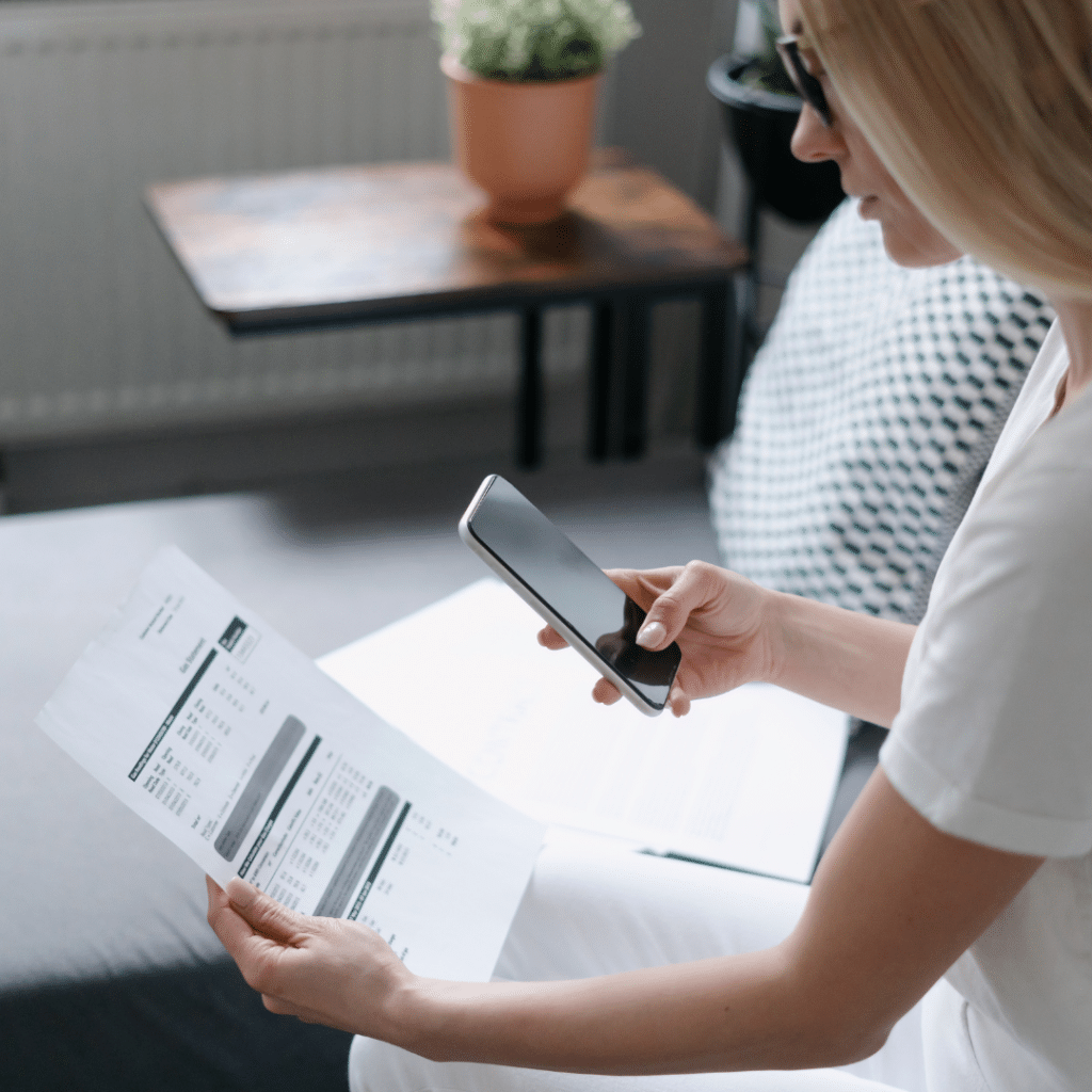 A woman sitting on a couch is using a smartphone to scan a document. She is wearing a white shirt and glasses, and a black and white patterned pillow is beside her. A table with a potted plant is in the background.