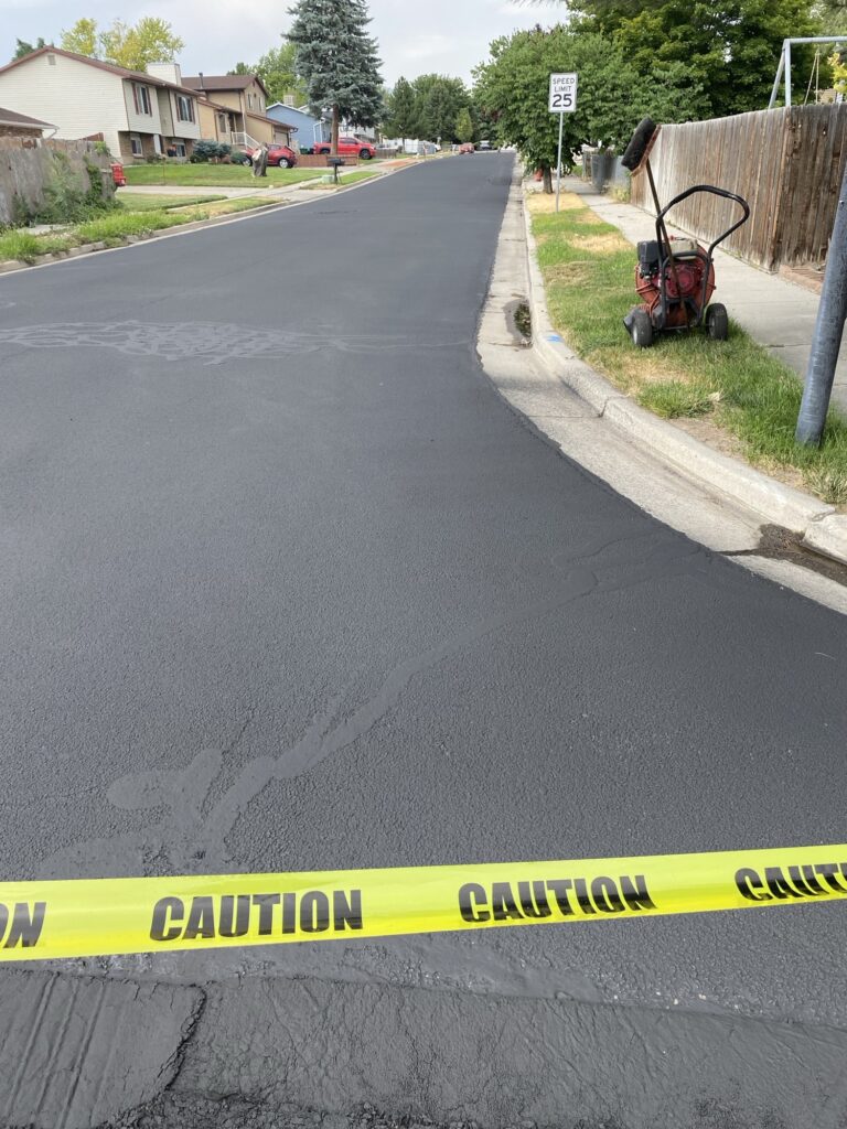 A freshly paved asphalt road with a yellow Caution tape across the entrance. A lawnmower is on the grass to the right, and houses line the street in the background. A speed limit sign reads 25 mph.