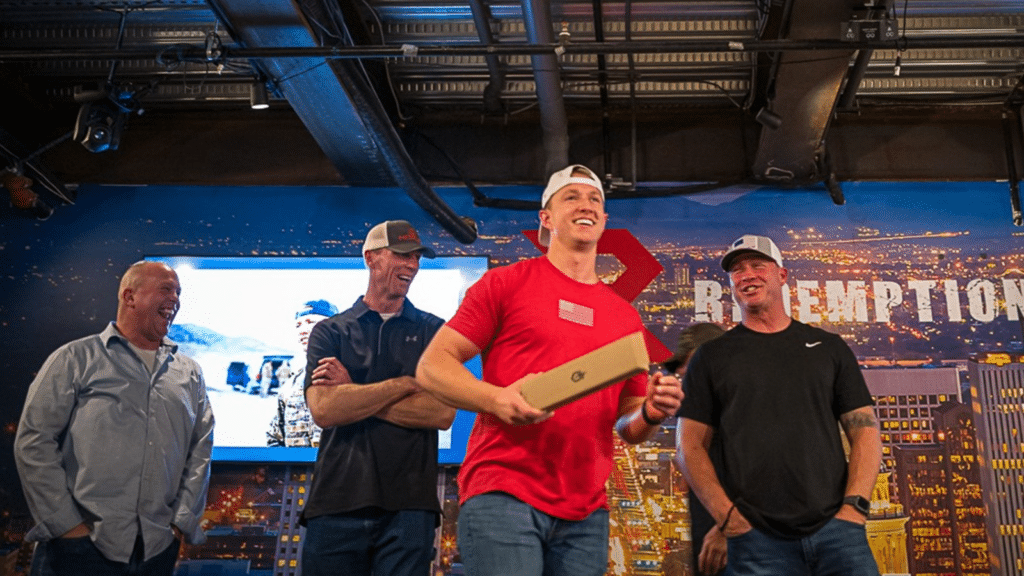 Four men are on stage, smiling and laughing. One in a red shirt holds an award. They stand in front of a city-themed backdrop with the word REDEMPTION. The atmosphere appears celebratory and informal.