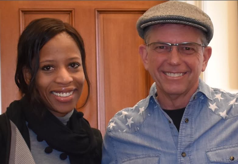 A woman with long dark hair and a black scarf smiles next to a man in a gray flat cap and glasses. He is wearing a blue button-up shirt with white stars on the shoulders. They are standing in front of a wooden door.