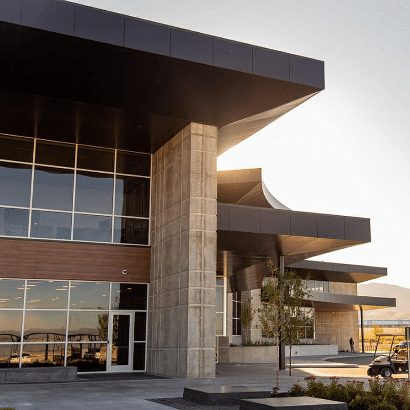 Modern building with large glass windows and overhanging roof, featuring concrete and wooden accents. Sunlight illuminates the structure from behind. The foreground has paved pathways and small trees.