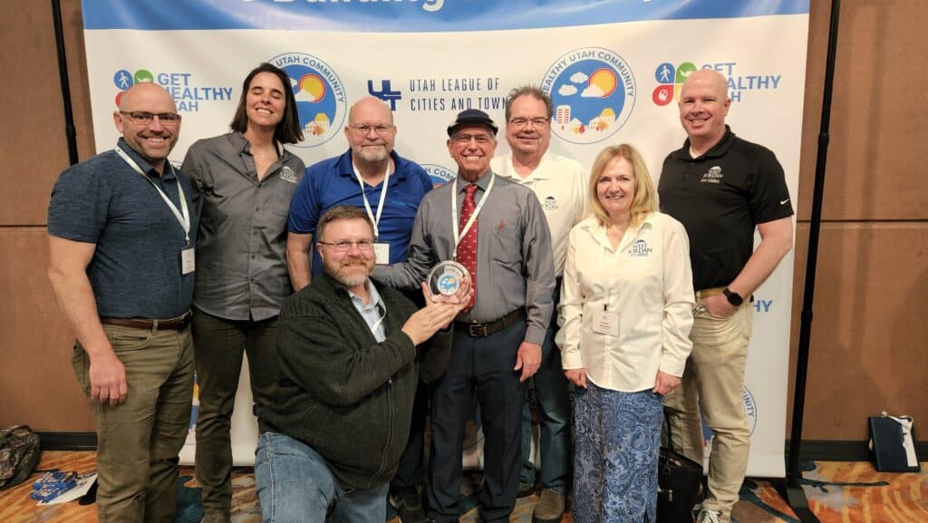 A group of eight adults, smiling and posing together at an indoor event. One person in the center holds a round award. A banner behind them displays Get Healthy Utah and other logos.