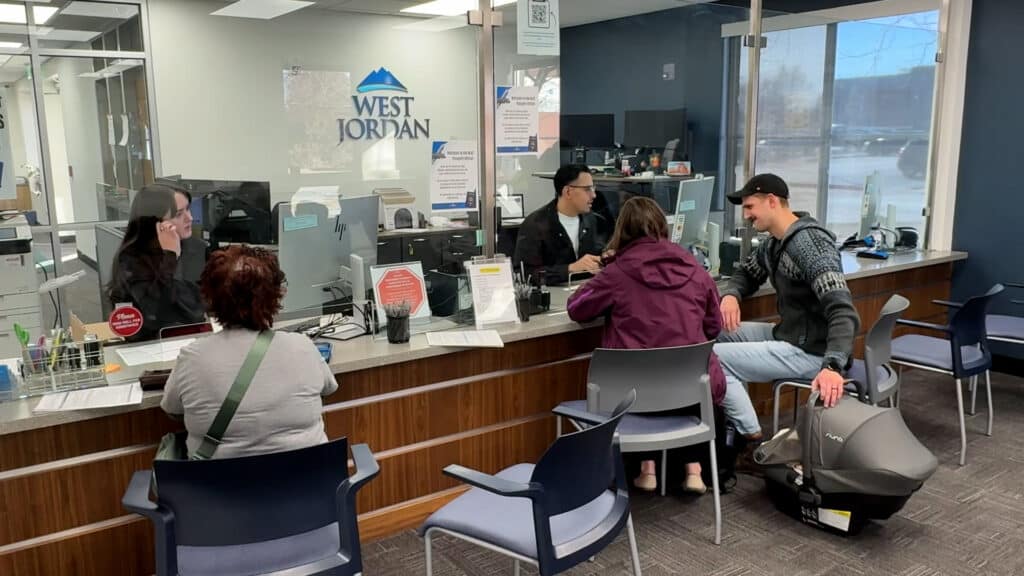 Several people sit at a service counter inside a municipal office with a West Jordan sign on the wall. Two staff members assist visitors through glass partitions, while others wait in chairs nearby.