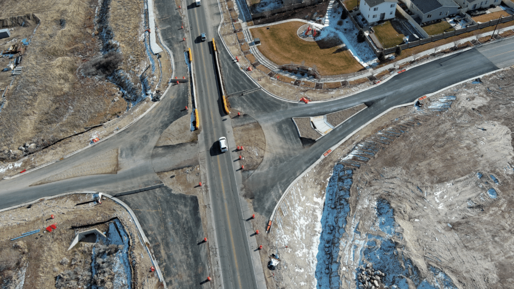 Aerial view of a road intersection under construction, with traffic cones and barriers placed along the roads. Several vehicles are navigating the intersection. Surrounding areas include residential houses, dirt, and patches of snow.