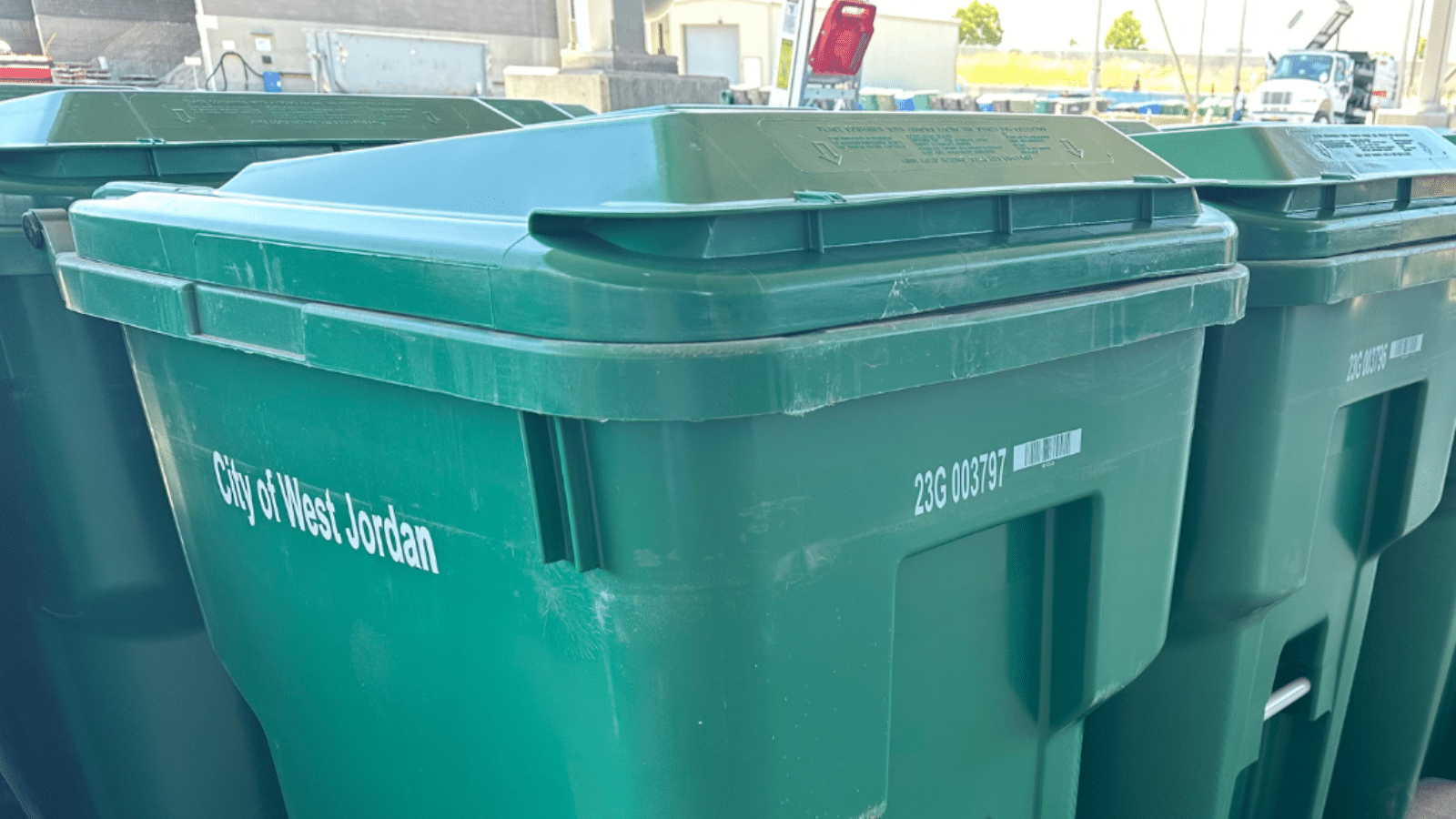 Green recycling bins with wheels are lined up in an outdoor area. The front bin is labeled City of West Jordan with a barcode and numerical code. Other bins and some buildings are visible in the background.