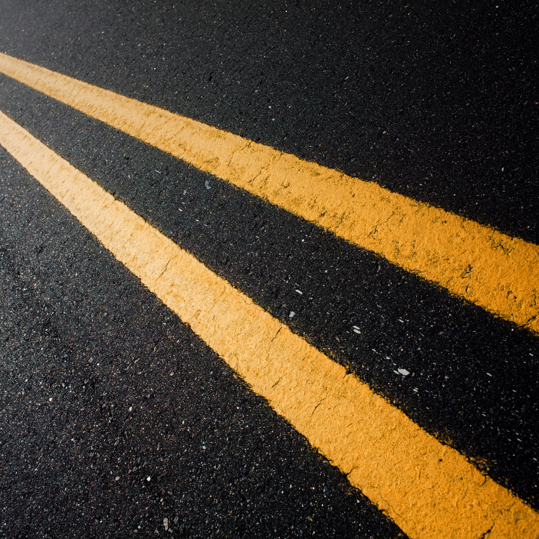 Close-up view of two parallel yellow lines on an asphalt road, painted diagonally across the image. The lines are slightly worn, indicating they are on a well-traveled road. The surface appears textured and dark.