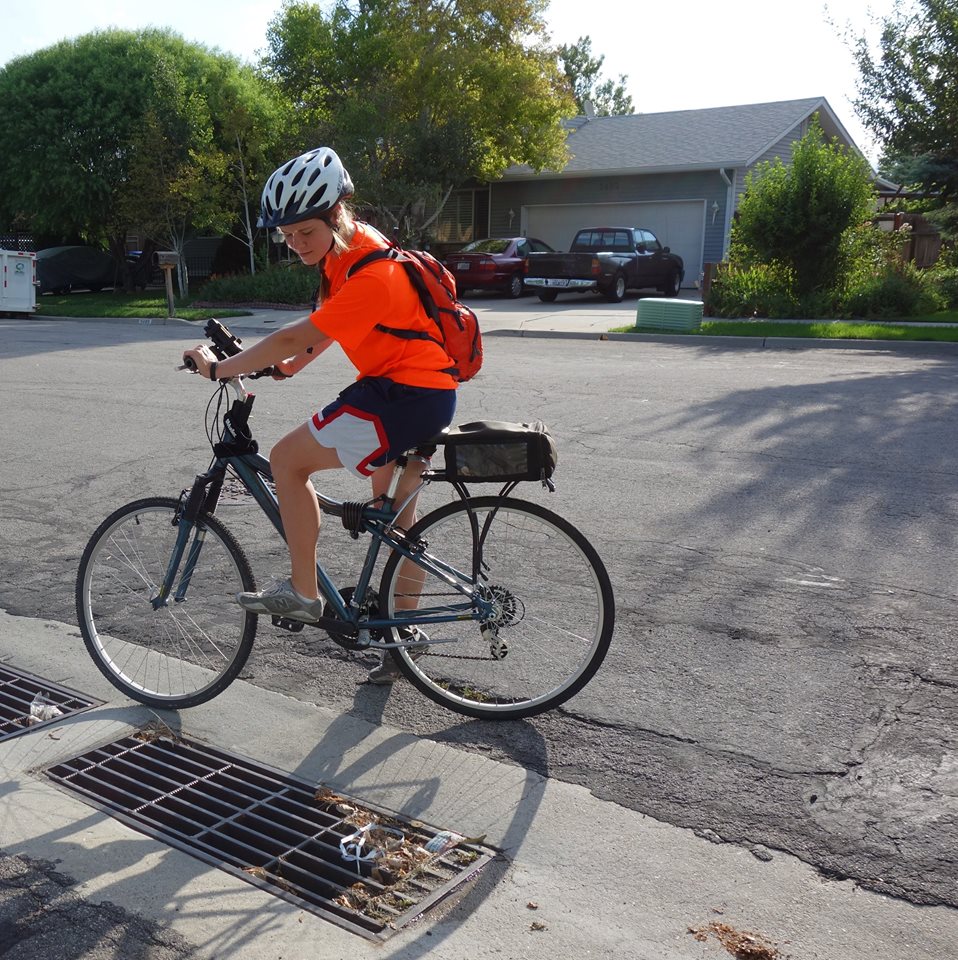 A young person wearing a helmet, orange shirt, and backpack rides a bicycle on a residential street, near a storm drain, on a sunny day. Houses and greenery are visible in the background.