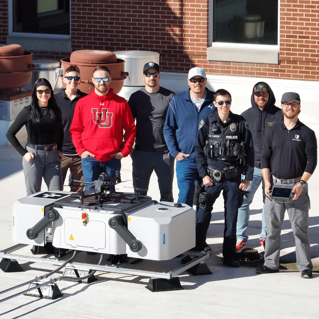 A group of eight people stand on a rooftop beside a large drone. One person wears a police vest, others in casual clothing and sunglasses. Theyre smiling, with a brick wall and equipment visible in the background.