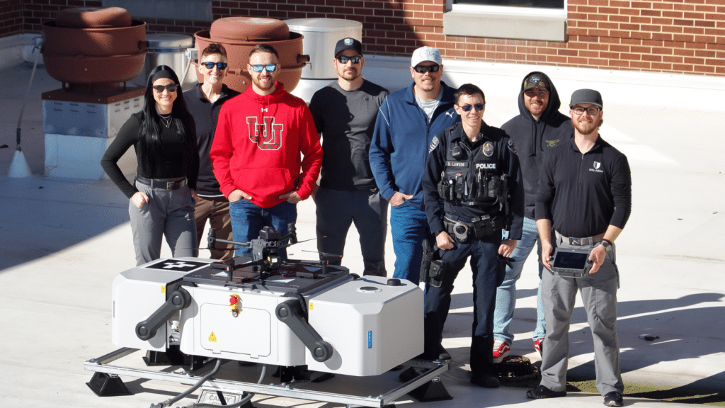 A group of seven people, including a police officer and a person with a tablet, stand outdoors on a rooftop with a large drone on a platform. Theyre smiling, with a brick building in the background.