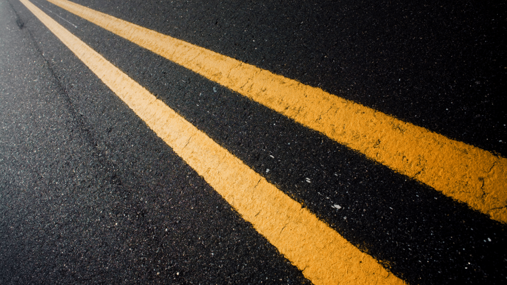 Close-up of two parallel yellow lines on a dark asphalt road, suggestive of road markings. The lines run diagonally across the image, showcasing texture and contrast between the vibrant yellow and the dark pavement.